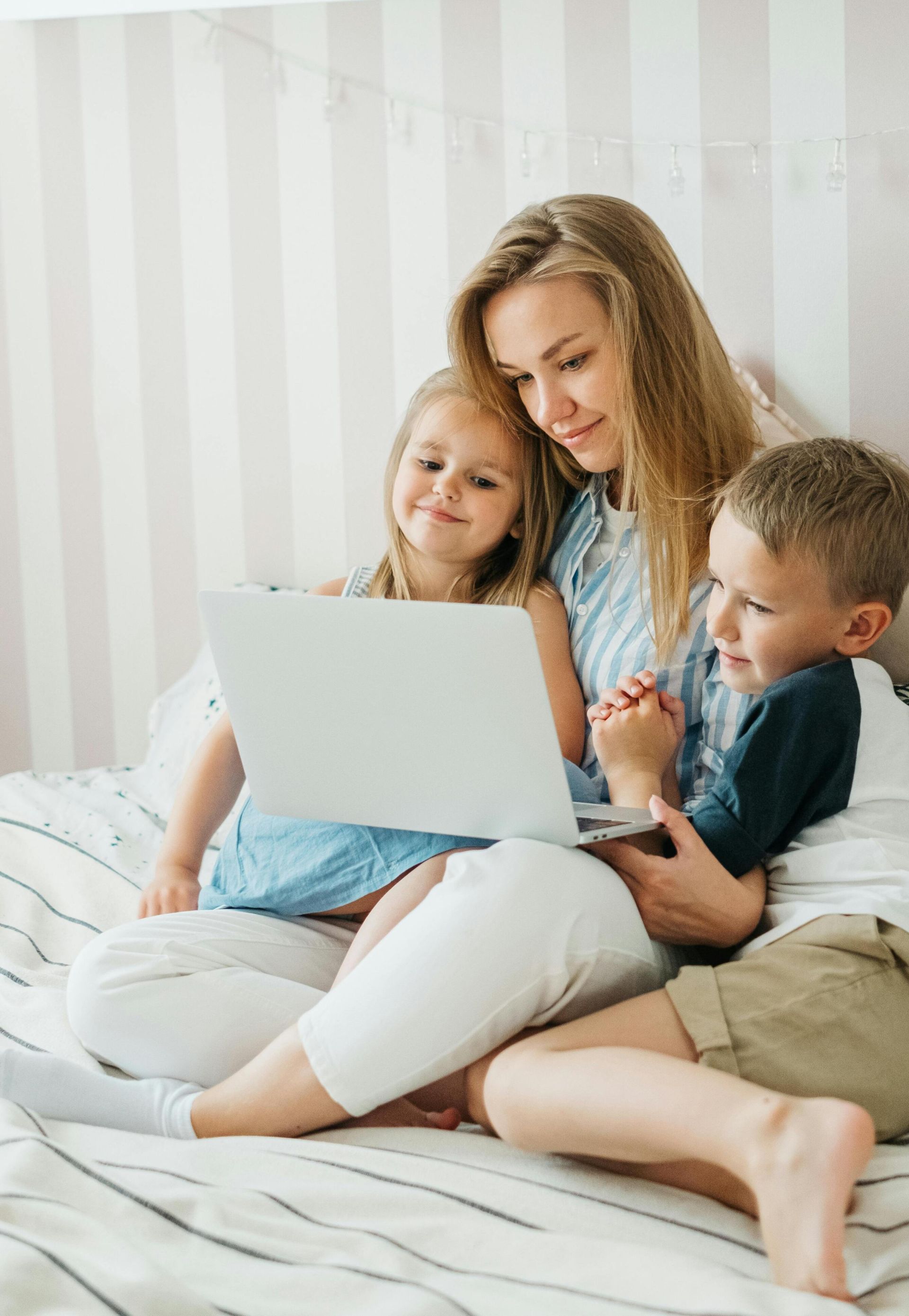 A mother sits on a bed with her children, sharing a joyful moment using a laptop.